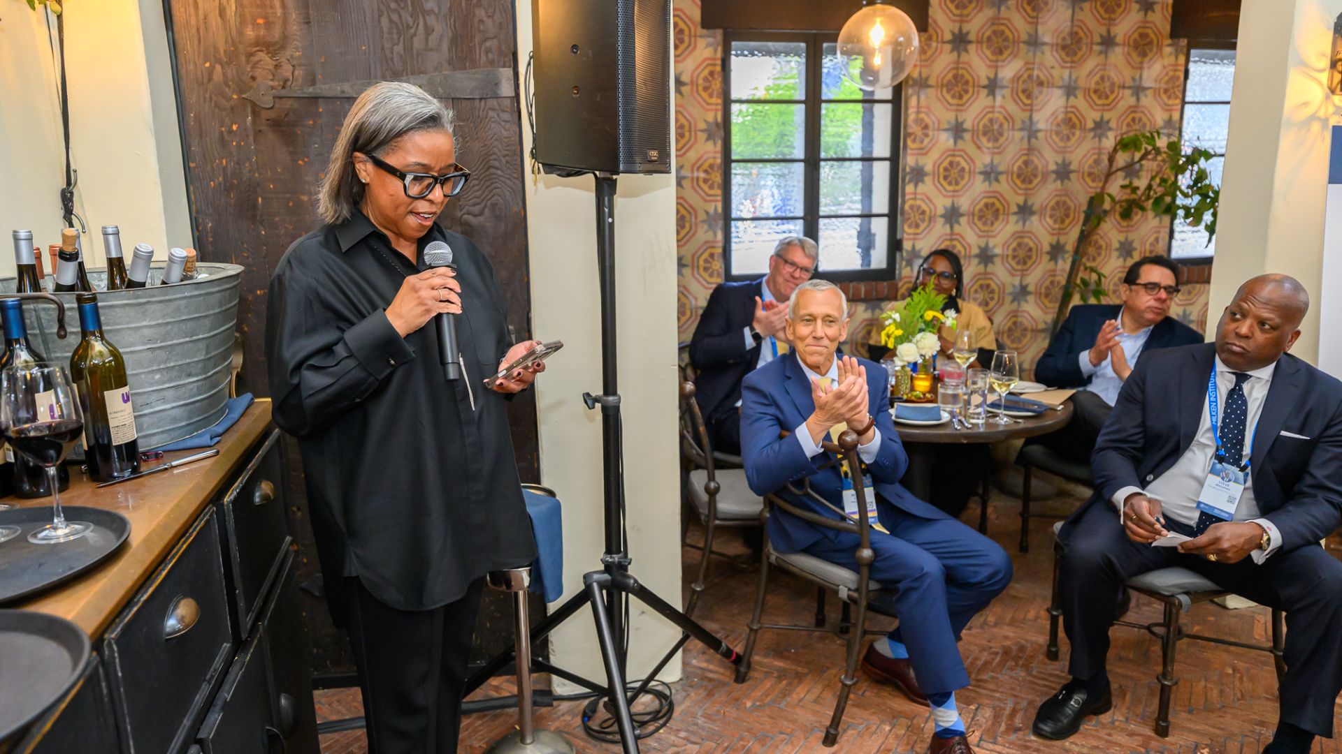 Woman speaking at microphone while three people listen seated in rustic interior setting